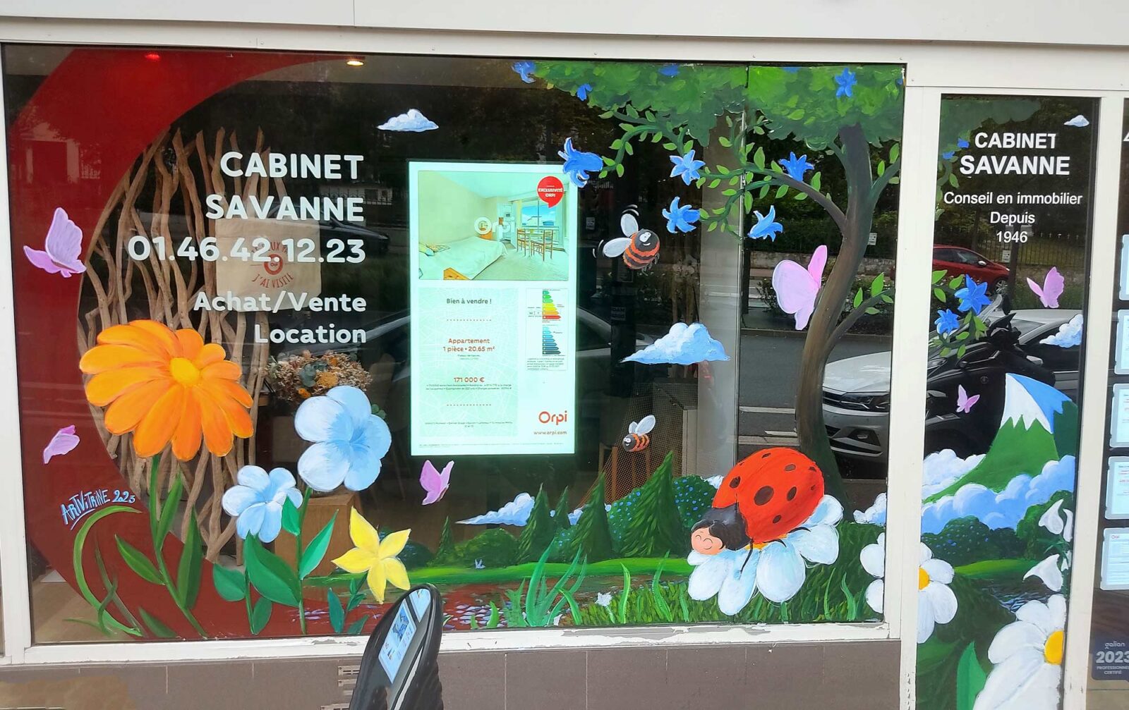 Peinture à la main sur vitrine pour une agence immobilière. La peinture représente un paysage printanier avec montagne coccinelle, abeilles et papillons (illustration). Agence orpi vanves 2023